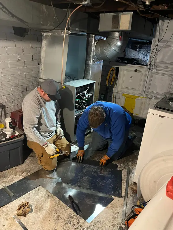 Two Affordable Air technicians cutting sheet metal for ductwork beside a furnace in a basement