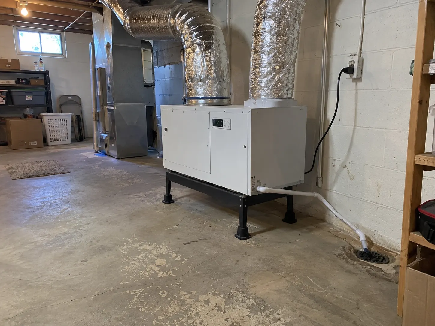 A whole-home dehumidifier installed on a stand in a basement, ducted to the HVAC system with a drain line to the floor drain