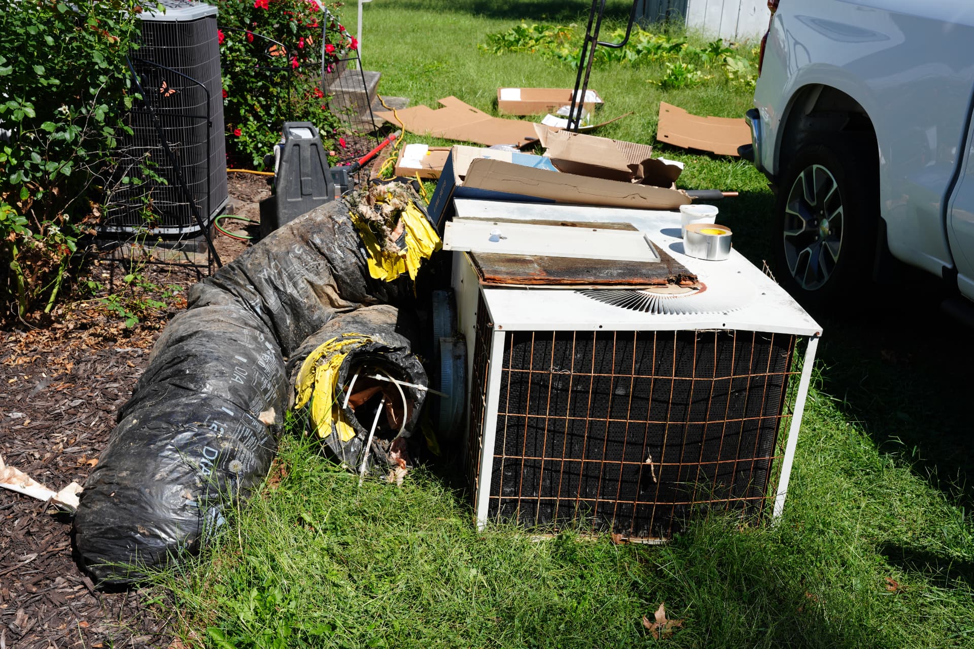 An old air conditioner and worn flexible ductwork removed during a replacement, beside the new outdoor unit